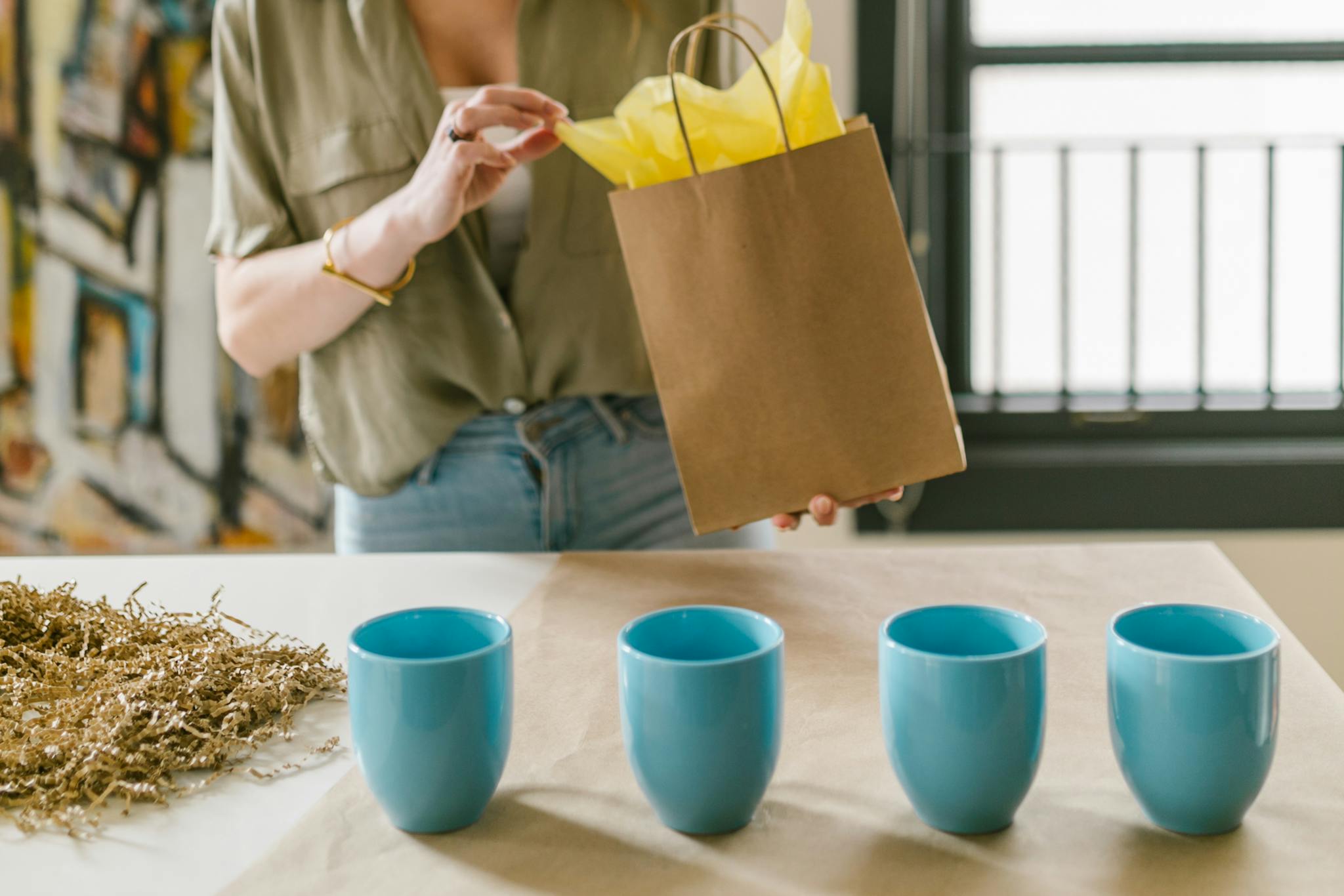 A woman packing a brown paper bag with colorful tissue alongside blue cups on a table.
